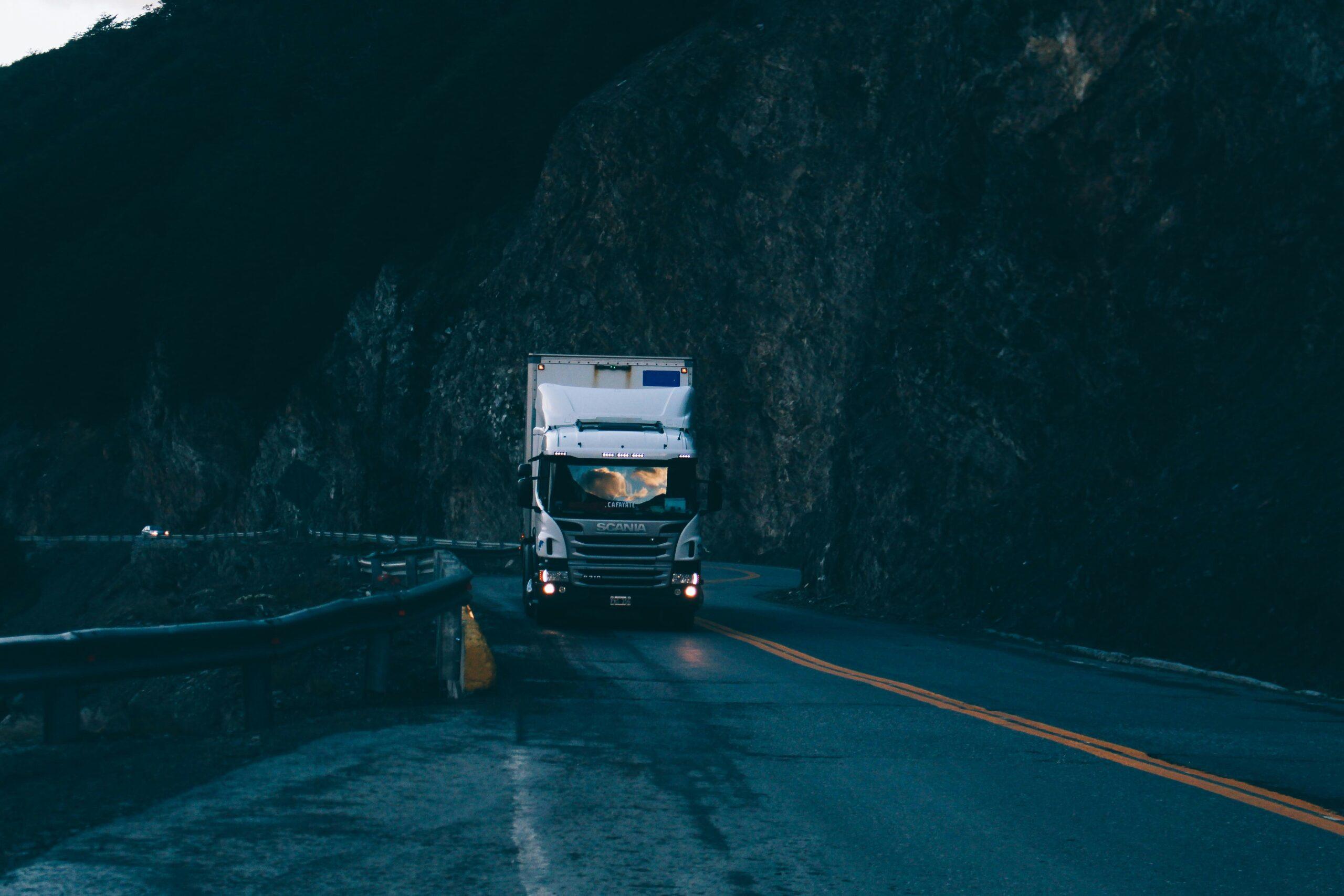 White semi-truck driving through a winding mountain road at dusk, headlights on, surrounded by dark rocky cliffs and low light conditions.