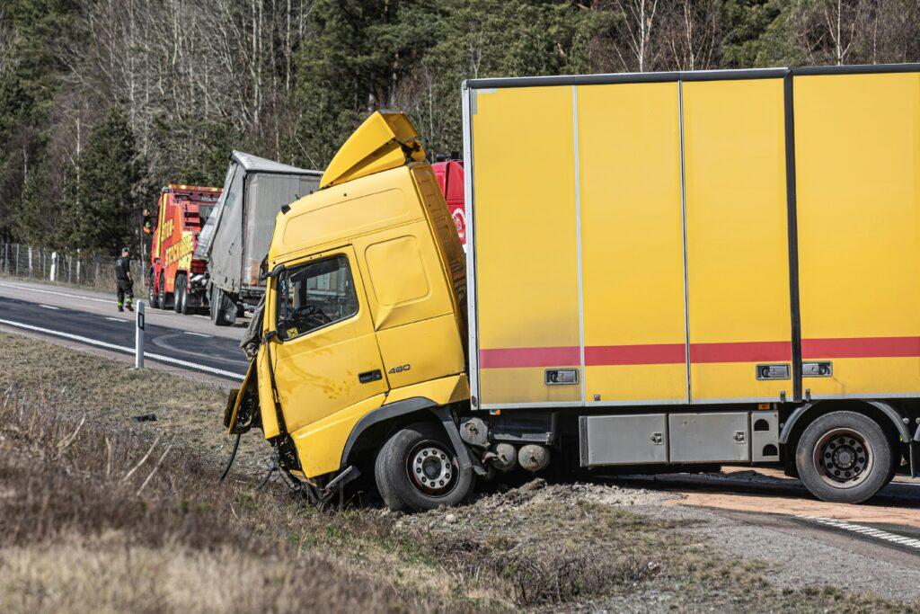 Yellow semi-truck crashed off the side of the road with visible front-end damage, rescue trucks in the background, and emergency personnel on scene.