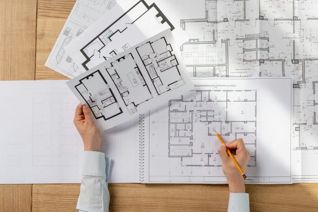 Hands reviewing detailed architectural blueprints spread across a desk, comparing floor plan layouts and marking notes with a pencil.