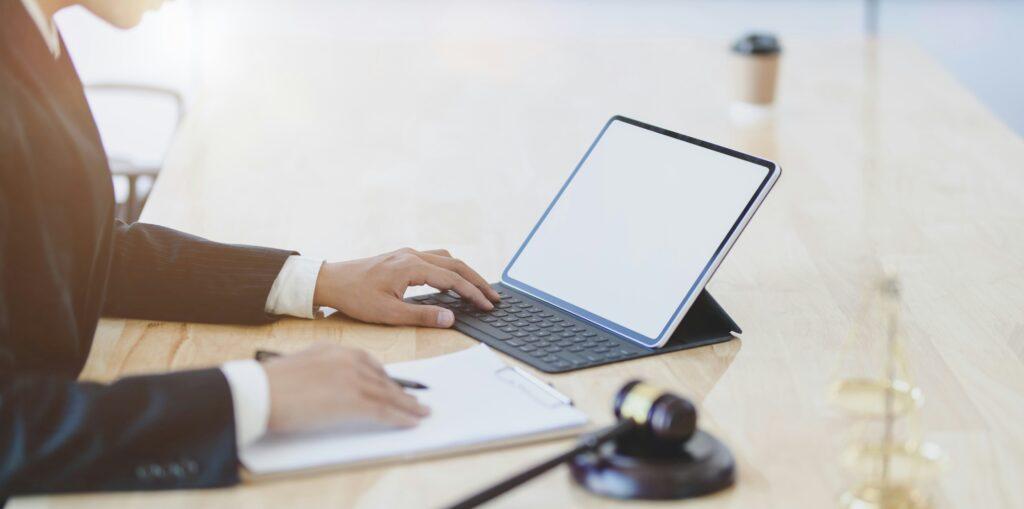 A lawyer sitting at a wooden desk using a tablet with a keyboard, with a clipboard, gavel, and blurred scales of justice in the foreground.