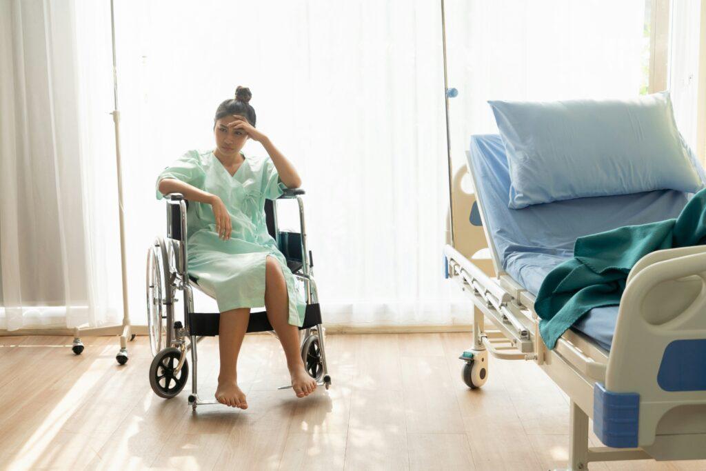 A young woman in a hospital gown sitting barefoot in a wheelchair beside an empty hospital bed, looking tired and distressed in a bright medical room.