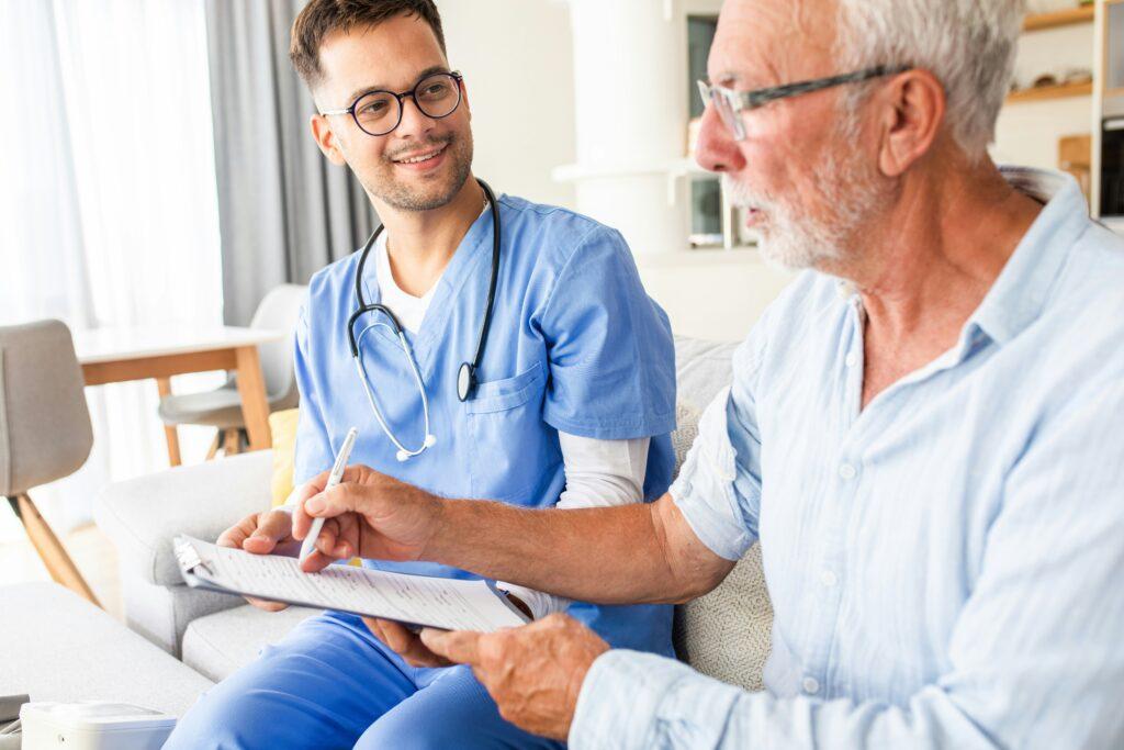 An elderly man reviewing paperwork on a clipboard with a smiling healthcare worker in scrubs sitting beside him in a bright home setting.