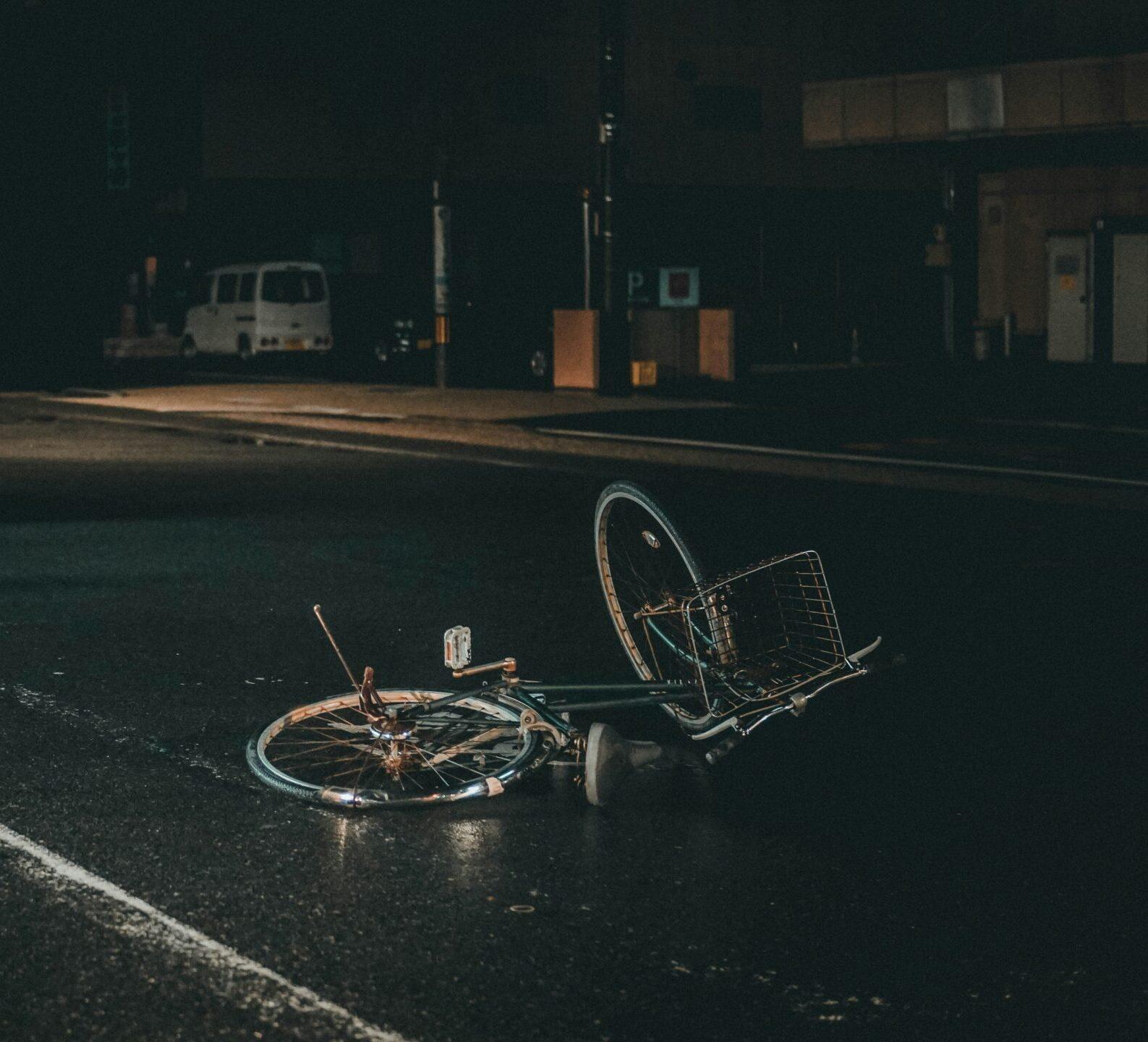 A damaged bicycle lying on its side in the middle of a dark, wet street at night, with a parked car and dimly lit buildings in the background.