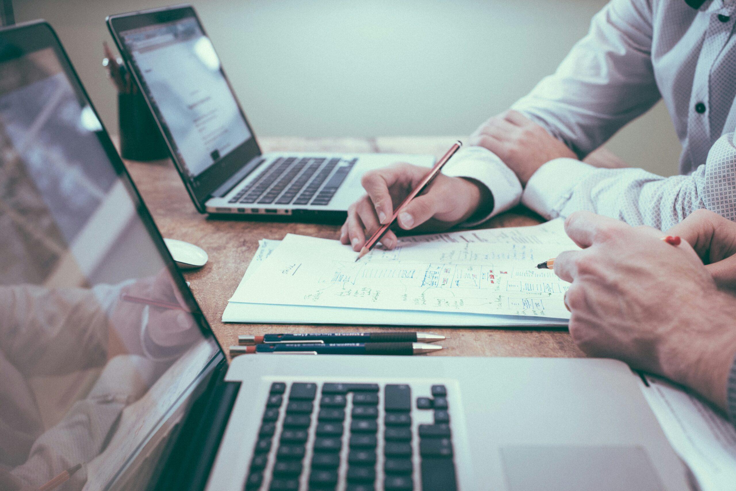Two people working at a desk with laptops and handwritten notes, reviewing documents together and discussing details.