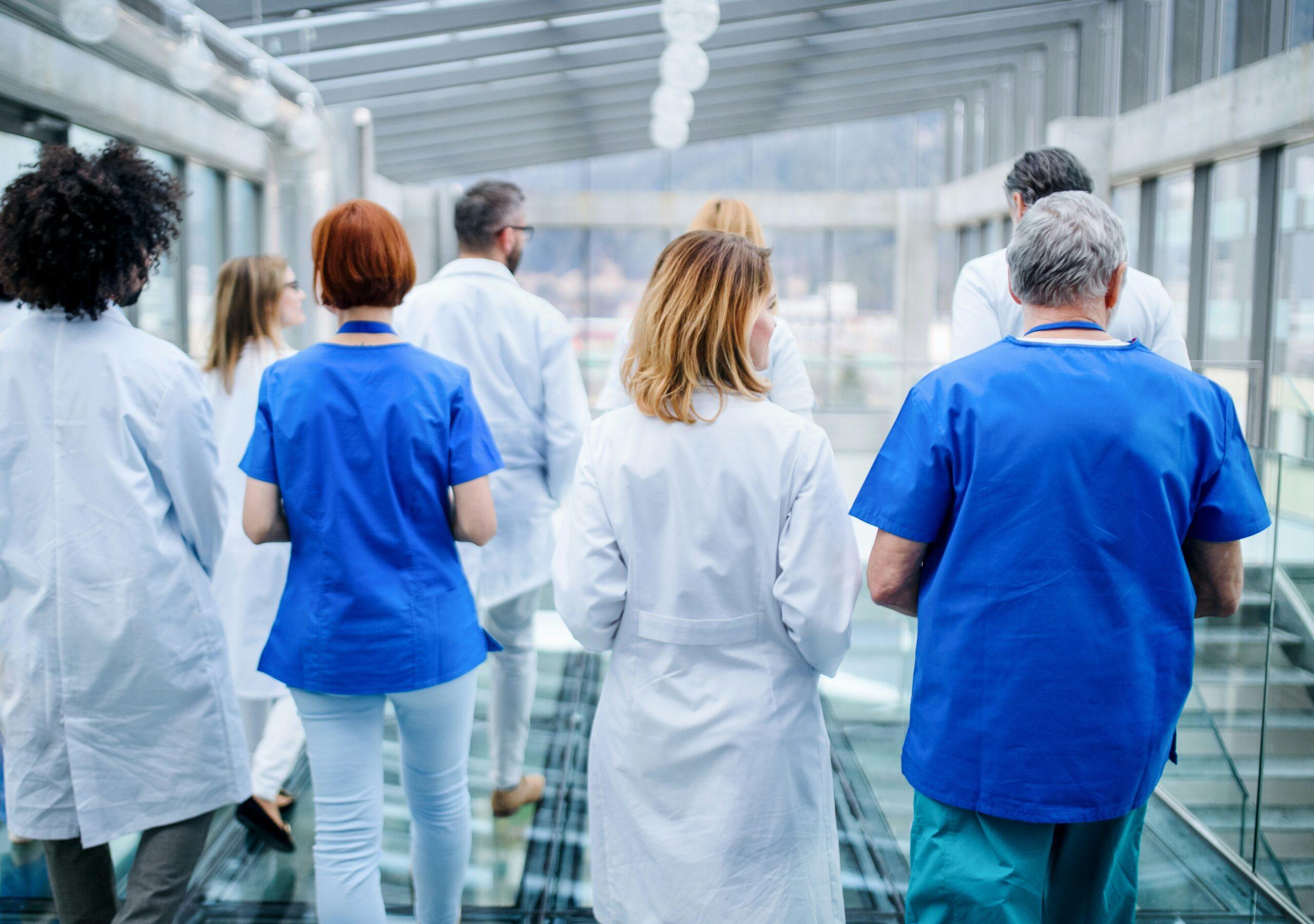 Eight medical professionals wearing assorted coats and scrubs are walking down a wide, brightly-lit area of a hospital. The POV is that they are walking away from you.