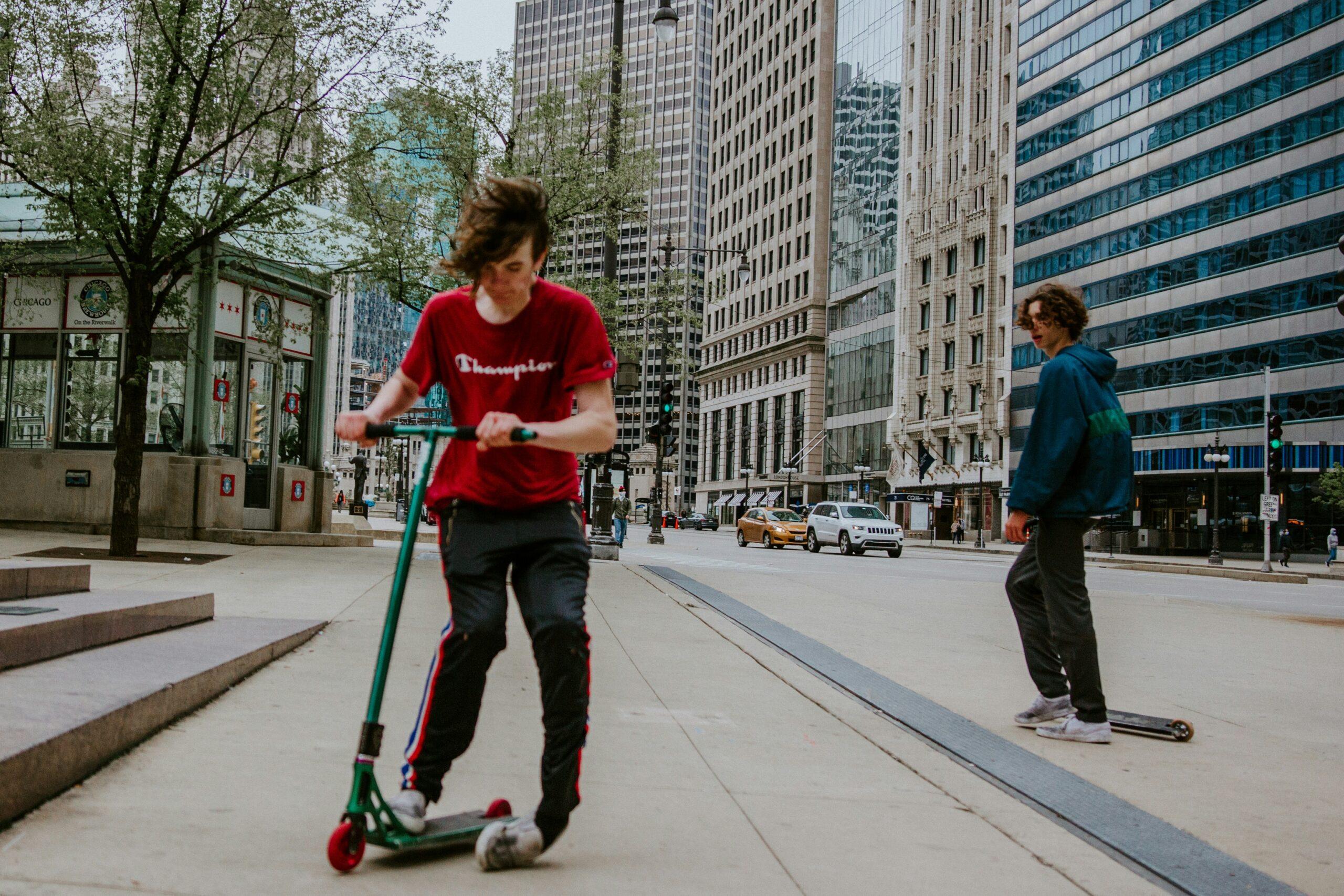 A teenaged boy with floppy brown hair and a red Champion-brand teeshirt is on a e-scooter; he appears to have rolled or broken his left ankle.