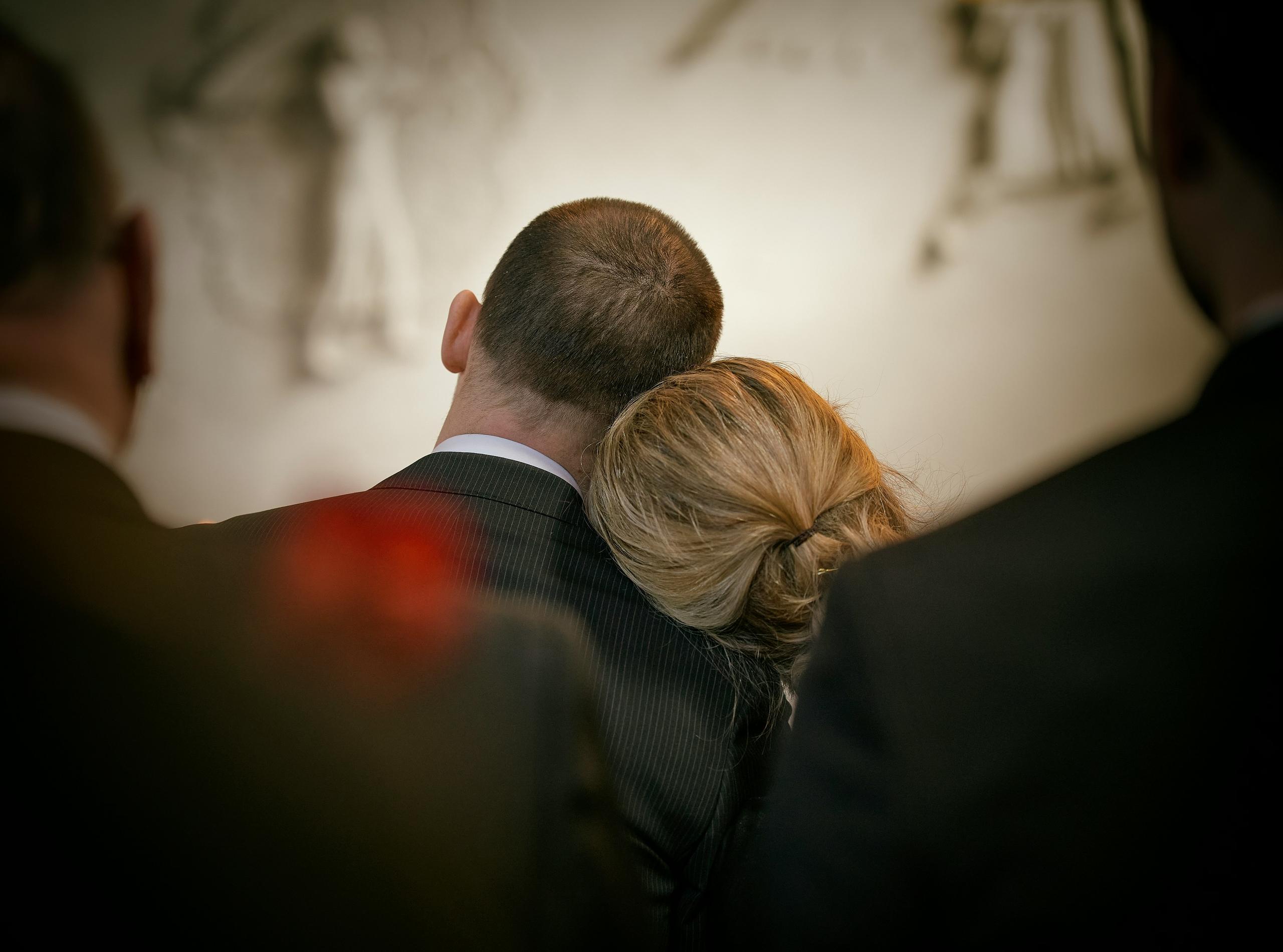 a woman with blonde hair leans on a man's shoulder. They are dressed in black for a funeral, and our POV is from behind them.