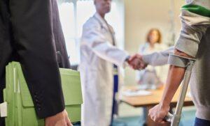 in the foreground are two men's arms: one is in a black suit and holding a thick green accordion file; the other is using a crutch. In he background are two medical professionals.