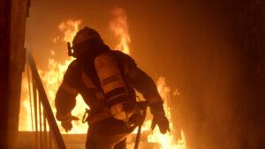 A firefighter walks through a burning building, navigating smoke and flames while carrying firefighting equipment.