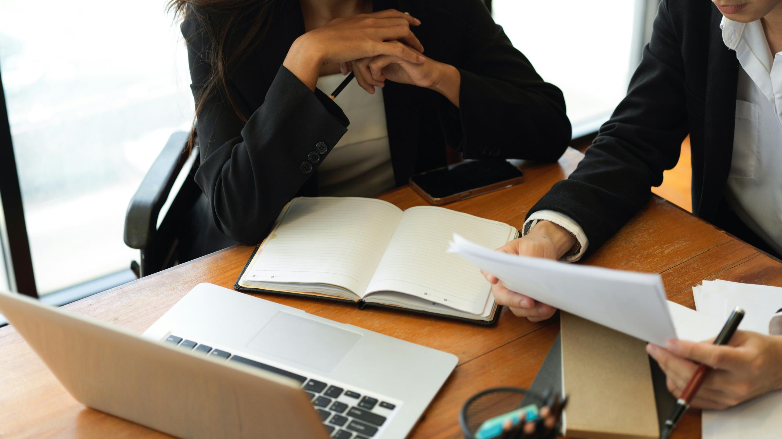 Two business professionals seated at a table, reviewing documents and working on a laptop.