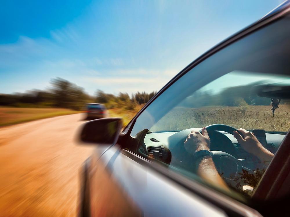 Person’s hands on the steering wheel of a car speeding down a rural road behind another vehicle