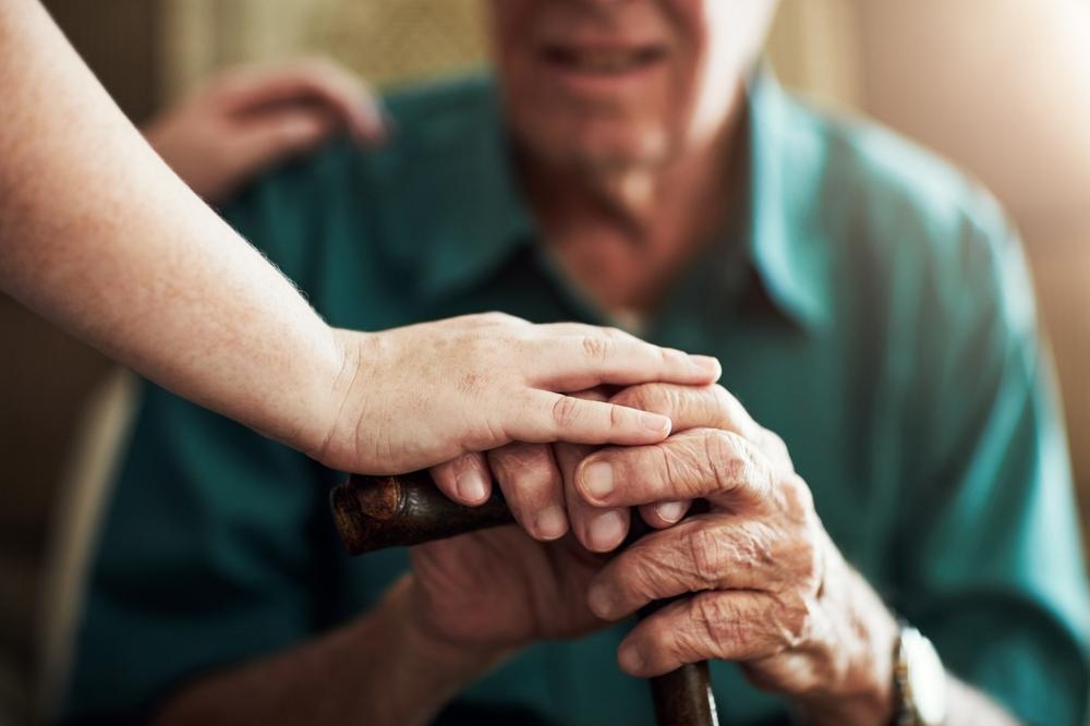 Person resting a comforting hand on an elderly man’s hands as he holds a cane