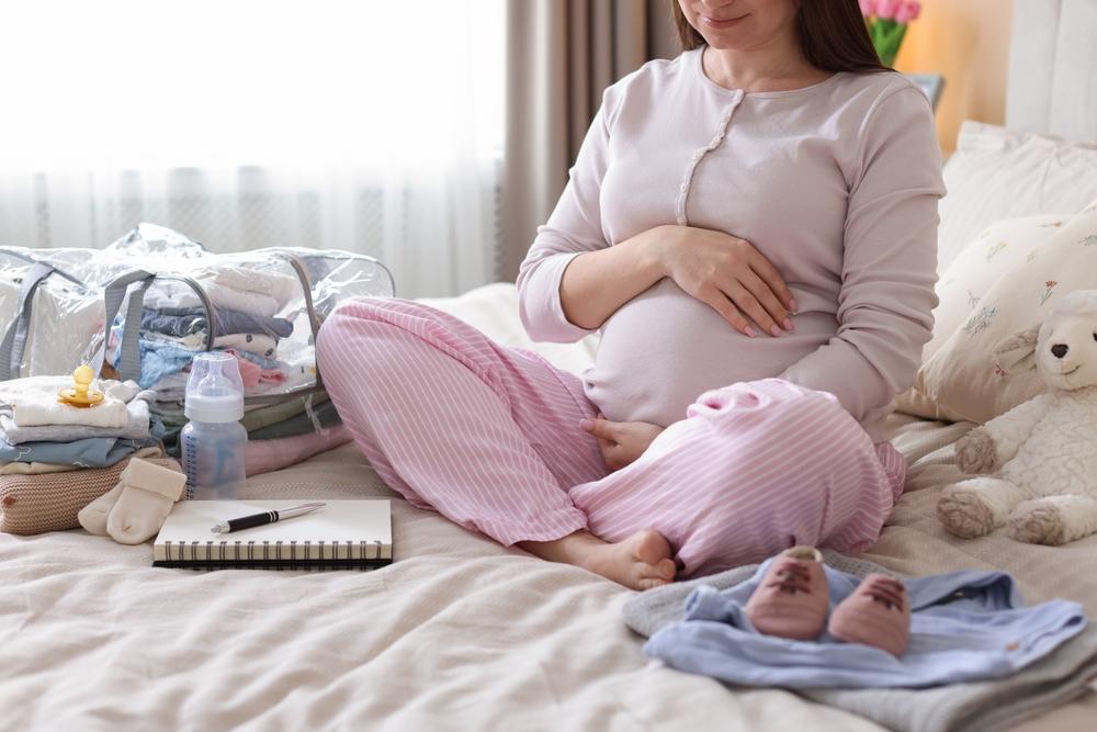 Pregnant woman sitting on a bed with a hand on her belly, beside a packed hospital bag, baby clothes, a bottle, a pacifier, baby shoes, and a notebook
