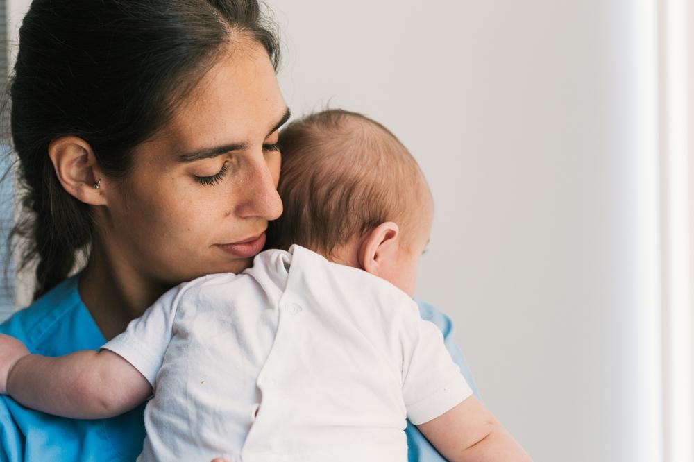 Woman in medical scrubs holding a baby against her shoulder, eyes closed and cheek resting on the baby’s head