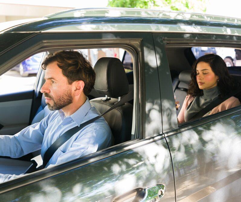 Man driving a car with a woman riding in the back seat, both wearing seat belts