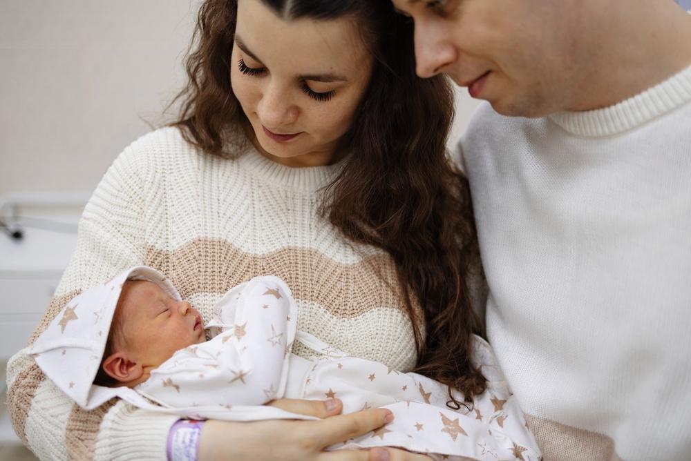 Woman and man smiling down at a sleeping newborn the woman holds in her arms, with a hospital wristband on her wrist